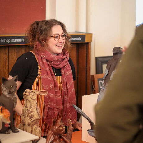 Middle aged white women with auburn hair tied up and a large brown scarf standing behind a craft stall