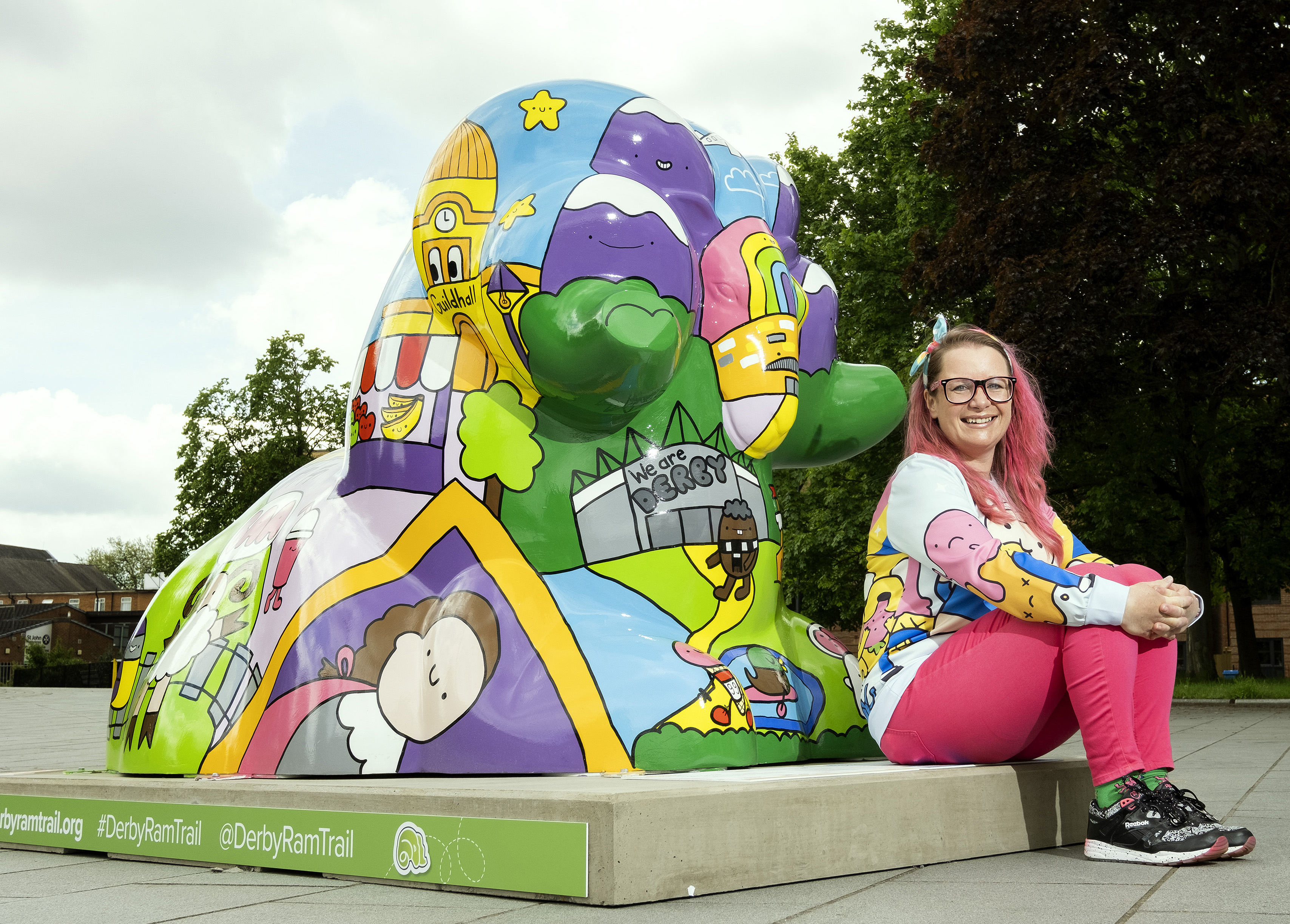 Middle aged white women with pink hair, cartoon style sweatshirt and pink trousers sits next to a colourful giant ram scuplture