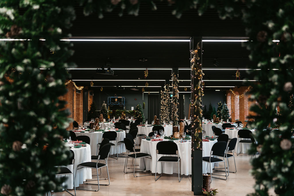 Spacious event hall decorated with white tablecloths and black chairs, adorned with festive greenery and string lights for a celebration.