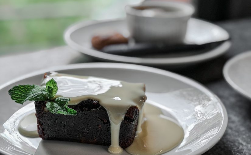 Traditional Christmas pudding served with a brandy sauce on a white plate with another dessert in the background in front of a window