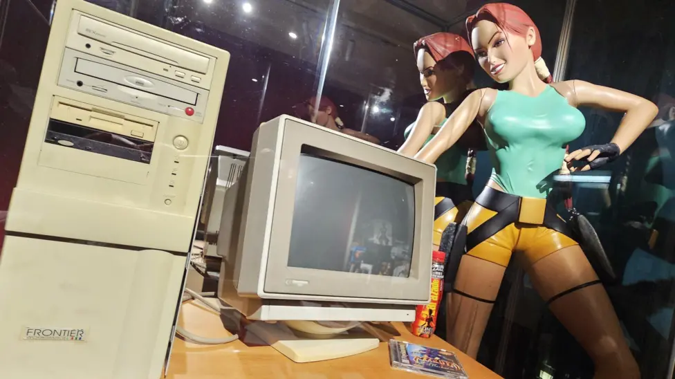Retro beige desktop computer setup with CRT monitor and tower next to a detailed Lara Croft action figure in a glass display.